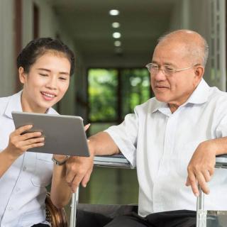A young woman assists an elderly man with a tablet device in a caregiving setting.