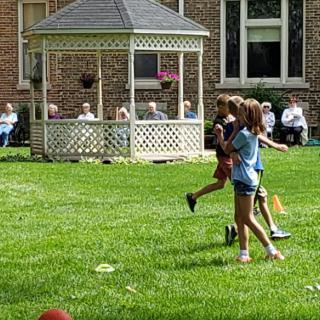 Children playing on a grassy lawn in front of a gazebo, with a group of people seated in the background.