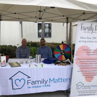 An outdoor booth for Family Matters In-Home Care, featuring two people at a table with informational materials and a colorful prize wheel.
