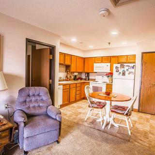 A cozy living room and kitchen area featuring a plush chair, dining table, and wooden cabinetry.