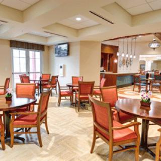 A well-lit dining area featuring wooden tables and red cushioned chairs, with a view of a bar in the background.