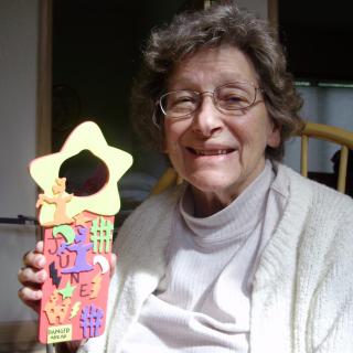 An elderly woman smiling while holding a colorful craft project.