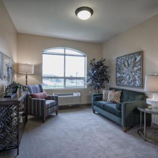 A cozy living room with a large window, decorated with a sofa, armchair, artwork, and lamps.