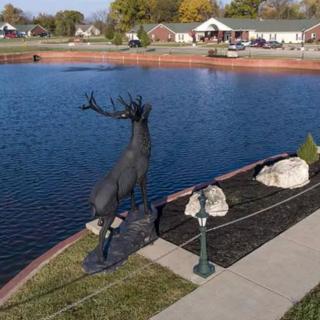 A statue of a deer stands beside a lake in a park setting.