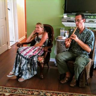 A man plays the flute while a woman listens in a comfortable living room setting.