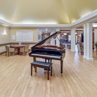 A spacious room with a grand piano at the center, surrounded by tables and chairs, featuring elegant lighting and wooden flooring.