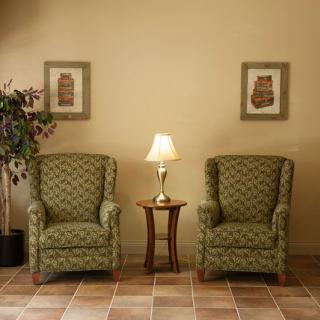 A cozy living room corner with two patterned armchairs flanking a small wooden table topped with a lamp, complemented by framed artwork on the wall and a potted plant in the background.