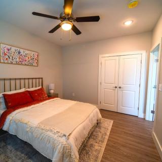 A cozy bedroom featuring a double bed with red pillows, a ceiling fan, and a nightstand with a lamp, all set against hardwood floors.