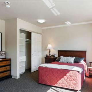 A cozy bedroom featuring a neatly made bed with a red cover, a wooden dresser, an open closet, and a bedside lamp.