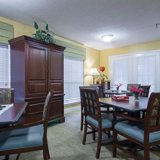 A cozy dining room with a wooden table and chairs, a large cabinet, and light streaming through windows with blinds.