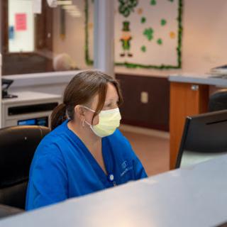 A healthcare worker in blue scrubs and a mask is focused on the computer in a hospital office.