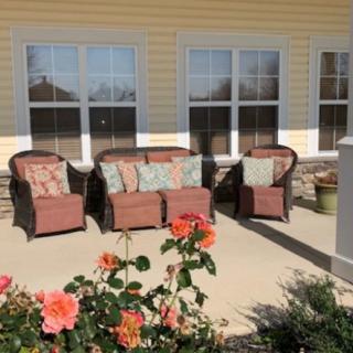A cozy outdoor patio area with chairs and a sofa, adorned with cushions, surrounded by blooming flowers.