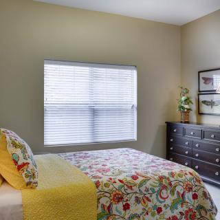 A cozy bedroom with floral bedding, a dark dresser, and natural light coming through a large window.