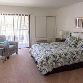 A cozy bedroom features a floral-patterned bed, an armchair by the window, and light streaming through the curtains.