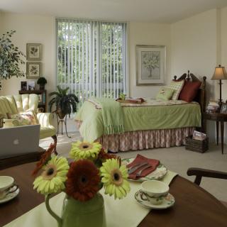 A cozy bedroom interior with a green color scheme, featuring a bed, natural light, and decorative plants and flowers.