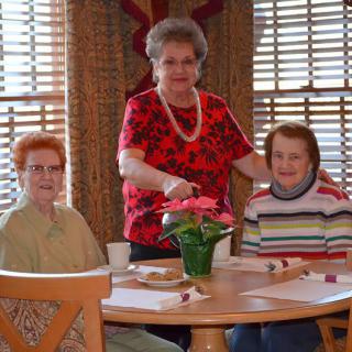 Three elderly women are gathered around a table indoors, with one standing and the other two seated. A flower pot is placed on the table.