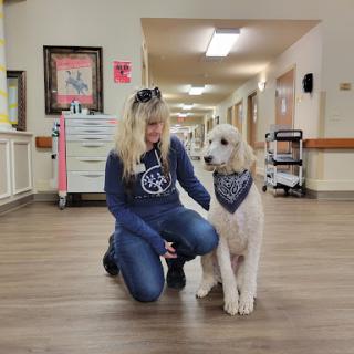 A woman kneels next to a friendly dog wearing a bandana in a brightly lit hallway.