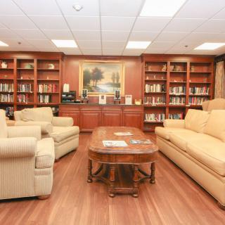 A cozy library room featuring bookshelves filled with books, comfortable sofas, a wooden coffee table, and a painting on the wall.