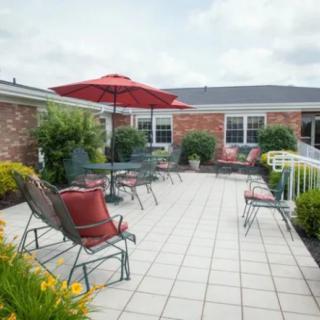 A spacious outdoor patio area with tables, chairs, and a red umbrella, surrounded by greenery and a brick building.
