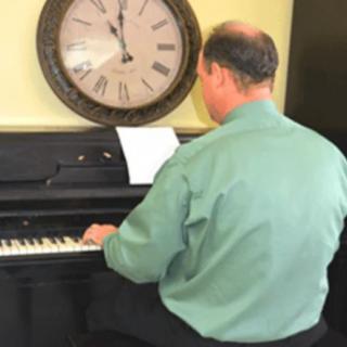 A man in a green shirt plays the piano beneath a clock on the wall.
