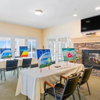 An art classroom setup with paintings displayed on easels, surrounded by tables and chairs, featuring a fireplace and television.