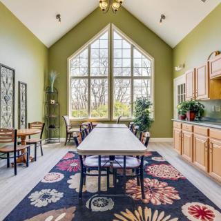 A spacious dining room with a high ceiling and large windows, featuring green walls, wooden furniture, and a decorative rug.