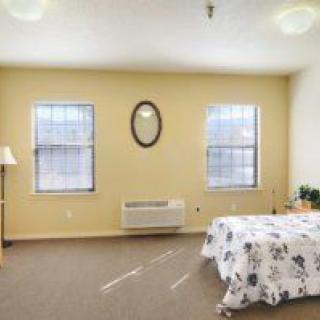 A simple, cozy bedroom featuring a bed with floral bedding, two windows, and an oval mirror on the wall.