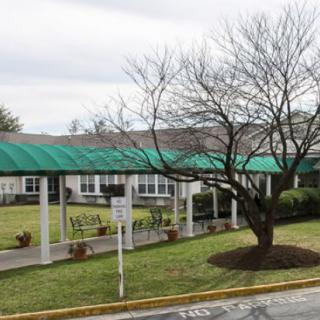 A building with a green canopy over a pathway, surrounded by a lawn and trees.