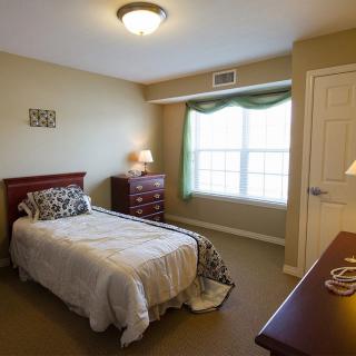 A cozy bedroom with a single bed, neatly arranged furniture, and natural lighting from a large window.