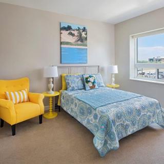 A brightly lit bedroom featuring a blue bedspread, a yellow chair, and a large window with natural light.