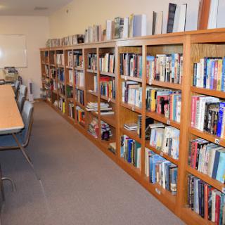 A cozy library space featuring wooden bookshelves filled with various books, accompanied by a long table and chairs for reading or studying.