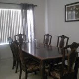 A dining room with a large wooden table and matching chairs, featuring a window with curtains and framed artwork on the wall.