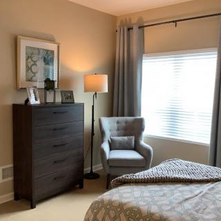 A cozy bedroom with a modern design featuring a dark dresser, an armchair, and a floor lamp by the window.