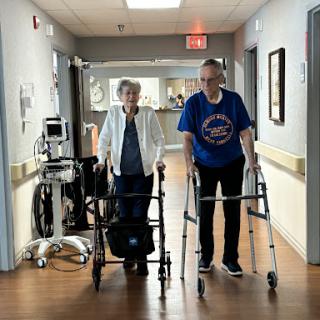 An elderly couple walking together with the help of walkers in a hospital corridor.
