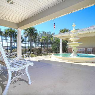 A sunny patio with a white bench and a fountain, surrounded by palm trees.