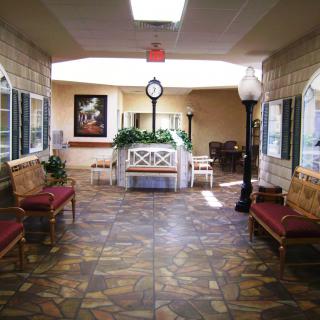 A spacious lobby area featuring decorative flooring, benches, artwork on the walls, and a central clock surrounded by plants.