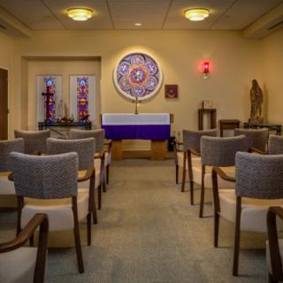 A quiet, intimate chapel with seating and decorative stained glass windows facing a central altar.