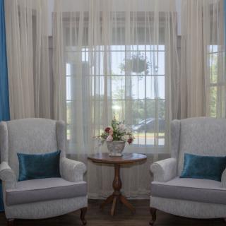 A cozy living room setting with two armchairs and a small table adorned with flowers, complemented by sheer curtains.