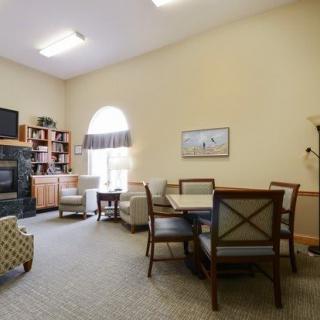 A cozy living room with comfortable chairs, a small table, bookshelves, and a television, illuminated by natural light from an arched window.