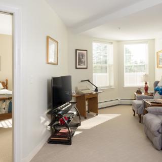 A cozy living room with natural light, featuring comfortable seating, a television, and a small desk.