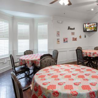 A cozy dining area with round tables covered in patterned tablecloths, surrounded by chairs and adorned with wall decorations. A television is mounted in the corner.