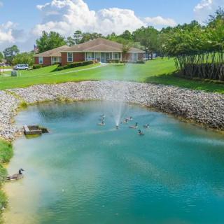 A serene landscape featuring a pond with a fountain in front of a house surrounded by grass and trees.