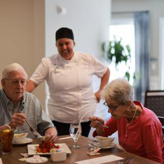 A chef engaging with two seniors while they enjoy a meal at a dining table.