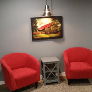 A cozy living room setup featuring two red chairs and a scenic painting illuminated by a lamp.