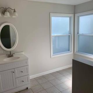A minimalist bathroom featuring a vanity with an oval mirror, tile flooring, and two large windows.
