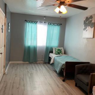 A minimalist bedroom with a single bed and chair, featuring teal curtains and a ceiling fan.