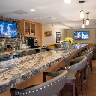 A modern indoor bar with a granite countertop, stylish chairs, and televisions.
