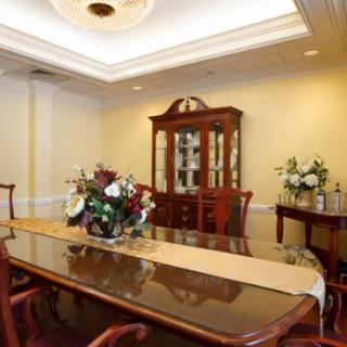 A formal dining room with a polished wooden table, surrounded by chairs, decorated with floral arrangements and a china cabinet in the background.