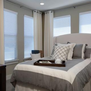 A cozy bedroom featuring a neatly made bed with decorative pillows and a tray, surrounded by large windows with blinds and curtains.