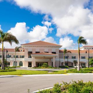 A large building with a red-tiled roof surrounded by palm trees and set against a backdrop of blue sky and clouds.